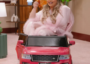 A person in a pink robe sits in a toy car, holding a slice of pizza, smiling. The room has a cozy, retro decor.