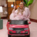 A person in a pink robe sits in a toy car, holding a slice of pizza, smiling. The room has a cozy, retro decor.