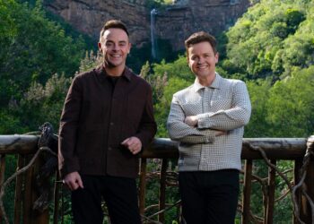 two individuals standing on a wooden balcony with lush greenery in the background