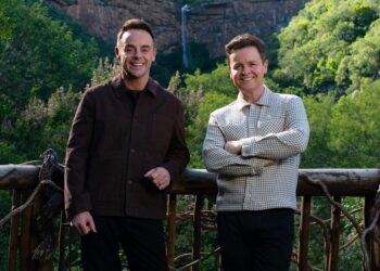 two individuals standing on a wooden balcony with lush greenery in the background