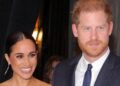 A smiling Meghan Markle stands next to Prince Harry, who wears a blue suit and tie with a white shirt.  She wears long gold-coloured earrings.