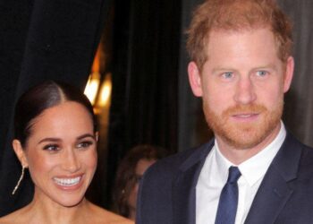 A smiling Meghan Markle stands next to Prince Harry, who wears a blue suit and tie with a white shirt.  She wears long gold-coloured earrings.