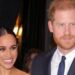 A smiling Meghan Markle stands next to Prince Harry, who wears a blue suit and tie with a white shirt.  She wears long gold-coloured earrings.