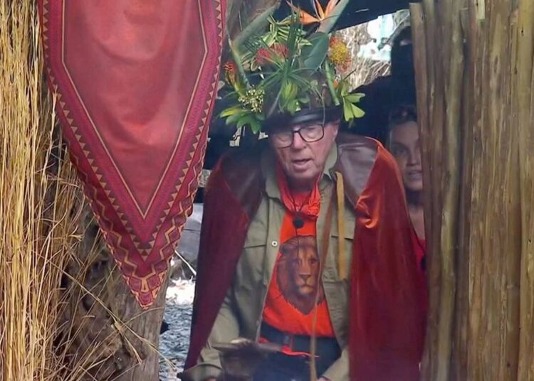 An elderly individual wearing a wreath of greenery and glasses is peering out from behind a red curtain, which appears to be part of a larger decorative setup.
