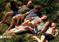 The Prince of Wales lies in the grass in a khaki tshirt and blue shorts, his hand is resting on his black spaniel dog, next to him is Prince George in a blue tshirt and green shorts, the Princess of Wales is smiling next to them in a white and navy stripped tshirt and white trousers, she holds the hand of Prince louis who wears a green shirt and chequered blue shorts, Princess charlottes is wearing black shorts and a pale green tshirt.