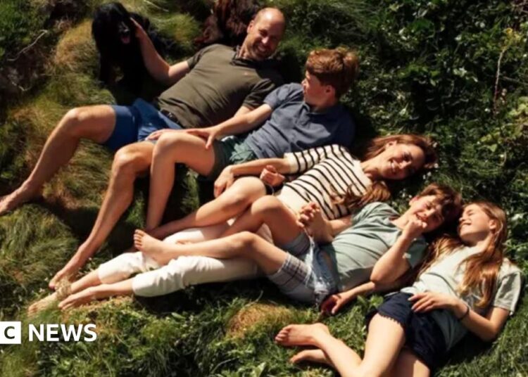 The Prince of Wales lies in the grass in a khaki tshirt and blue shorts, his hand is resting on his black spaniel dog, next to him is Prince George in a blue tshirt and green shorts, the Princess of Wales is smiling next to them in a white and navy stripped tshirt and white trousers, she holds the hand of Prince louis who wears a green shirt and chequered blue shorts, Princess charlottes is wearing black shorts and a pale green tshirt.