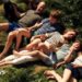 The Prince of Wales lies in the grass in a khaki tshirt and blue shorts, his hand is resting on his black spaniel dog, next to him is Prince George in a blue tshirt and green shorts, the Princess of Wales is smiling next to them in a white and navy stripped tshirt and white trousers, she holds the hand of Prince louis who wears a green shirt and chequered blue shorts, Princess charlottes is wearing black shorts and a pale green tshirt.