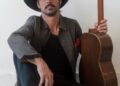 Ryan Bingham, wearing black cowboy hat and Western boots, sits on floor holding his guitar. (Elias Tahan photo)