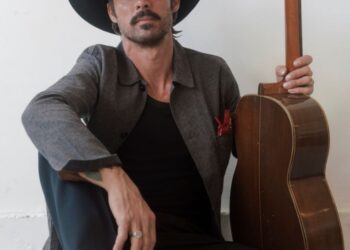 Ryan Bingham, wearing black cowboy hat and Western boots, sits on floor holding his guitar. (Elias Tahan photo)