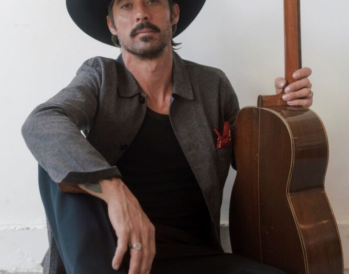 Ryan Bingham, wearing black cowboy hat and Western boots, sits on floor holding his guitar. (Elias Tahan photo)