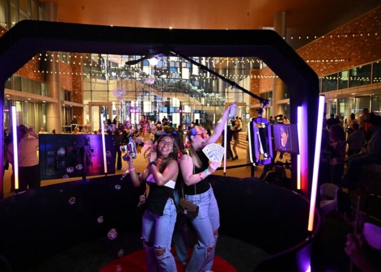 Two people pose and smile inside a photo booth setup by Meridian Events, surrounded by bubbles and bright lights, at a lively indoor gathering with a crowd in the background.