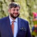 Eugene Mirman, wearing a blue suit, orange tie, and pocket square, smiles at the camera.