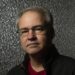 Vince Zampella wearing glasses, a dark jacket and red shirt poses in front of a dark backdrop