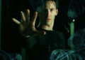 A man in a black shirt stands with his arm extended, palm out, appearing to stop or control swirling, translucent, blue-tinted objects frozen in midair around him. The scene has a dark, dramatic atmosphere.