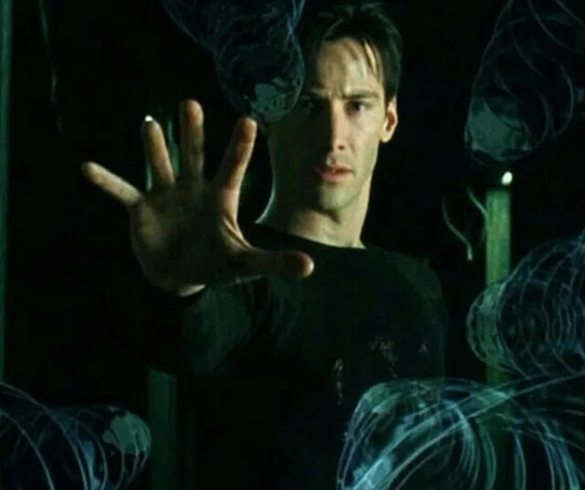 A man in a black shirt stands with his arm extended, palm out, appearing to stop or control swirling, translucent, blue-tinted objects frozen in midair around him. The scene has a dark, dramatic atmosphere.