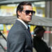 A person in a formal suit with a serious expression stands amid a crowd, appearing focused