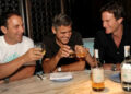 Three men sit at a marble table, smiling and toasting with glasses of liquor. There are two bottles on the table, and the setting appears to be a restaurant or bar with dim lighting and wine storage in the background.