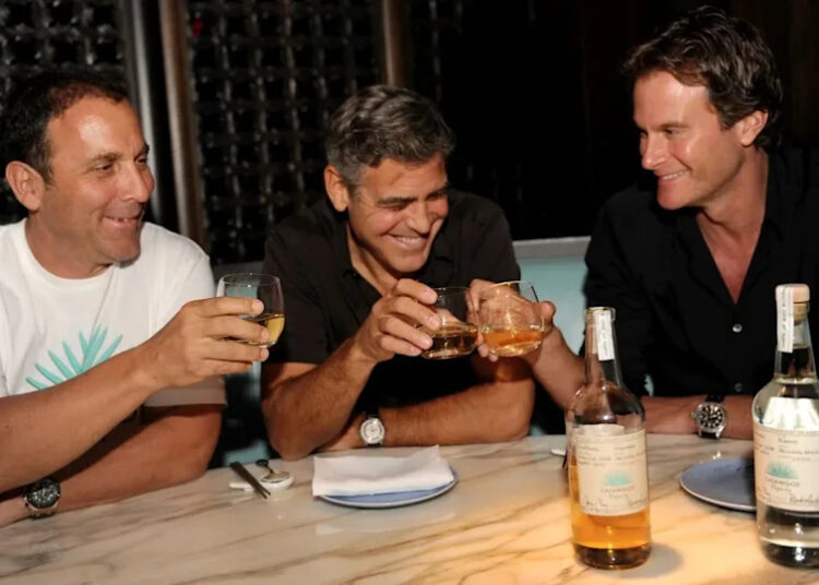 Three men sit at a marble table, smiling and toasting with glasses of liquor. There are two bottles on the table, and the setting appears to be a restaurant or bar with dim lighting and wine storage in the background.