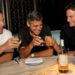 Three men sit at a marble table, smiling and toasting with glasses of liquor. There are two bottles on the table, and the setting appears to be a restaurant or bar with dim lighting and wine storage in the background.