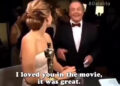 A woman in a white dress holding an Oscar statue talks to a smiling man in a tuxedo. The caption reads, "I loved you in the movie, it was great." A crowd and lights are visible in the background.
