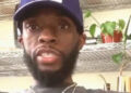 A man with a beard wearing a blue baseball cap with the number 42, a dark shirt, and necklaces sits indoors in front of metal shelves with plants and various items.