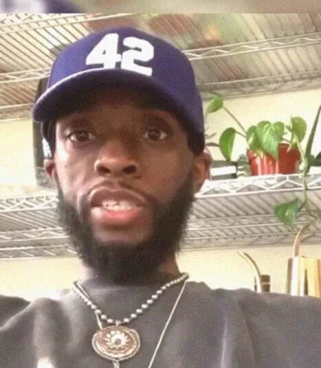A man with a beard wearing a blue baseball cap with the number 42, a dark shirt, and necklaces sits indoors in front of metal shelves with plants and various items.