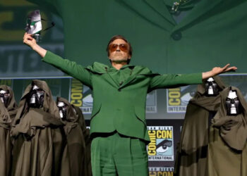 A man in a green suit stands center stage with arms outstretched, surrounded by people in dark cloaks and metal masks. He holds a mask in one hand. Comic-Con International logos are visible in the background.