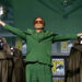 A man in a green suit stands center stage with arms outstretched, surrounded by people in dark cloaks and metal masks. He holds a mask in one hand. Comic-Con International logos are visible in the background.