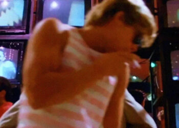 A person in a sleeveless striped shirt dances in front of a wall of old televisions showing various images, including a man in a suit. The scene is colorful and has a retro, 1980s vibe.