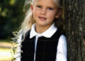 A young girl with long blonde hair, wearing a white collared shirt and black vest, stands outdoors leaning against a tree while holding a small bunch of white wildflowers. She looks at the camera and smiles slightly.