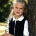 A young girl with long blonde hair, wearing a white collared shirt and black vest, stands outdoors leaning against a tree while holding a small bunch of white wildflowers. She looks at the camera and smiles slightly.