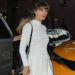 A woman with light brown hair and bangs wears a long-sleeved white ribbed top and a matching white pleated skirt, standing next to a yellow car on a city street. She carries a black purse and looks toward the camera.