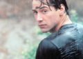 A young man with wet dark hair and a soaked black t-shirt looks over his shoulder in the rain, with a blurred natural background behind him.