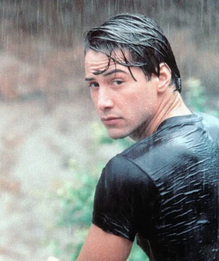A young man with wet dark hair and a soaked black t-shirt looks over his shoulder in the rain, with a blurred natural background behind him.
