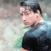 A young man with wet dark hair and a soaked black t-shirt looks over his shoulder in the rain, with a blurred natural background behind him.