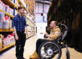 A man in a wheelchair talks with a standing man in a plaid shirt inside a warehouse lined with shelves full of labeled boxes and containers.