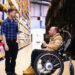 A man in a wheelchair talks with a standing man in a plaid shirt inside a warehouse lined with shelves full of labeled boxes and containers.