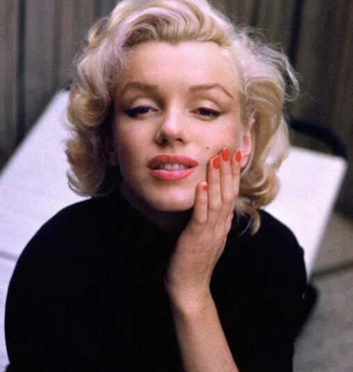 A woman with short, wavy blonde hair and red lipstick rests her hand on her cheek, wearing a black top and red nail polish, looking slightly up with a gentle expression. A blurred background is visible.