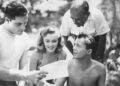 Four people gather, examining a paper, in a sunny outdoor setting with blurred foliage in the background.