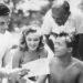 Four people gather, examining a paper, in a sunny outdoor setting with blurred foliage in the background.