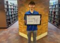 Student standing in a library holding a framed award certificate in front of a brick wall.