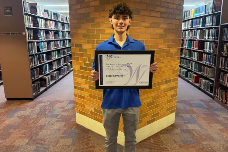 Student standing in a library holding a framed award certificate in front of a brick wall.