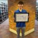 Student standing in a library holding a framed award certificate in front of a brick wall.