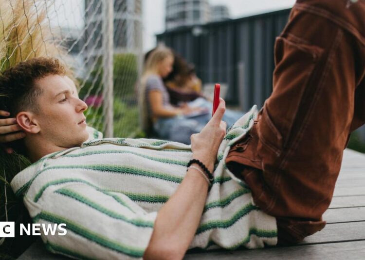 A young man lying down on the ground holding and scrolling on his smartphone.