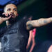 Drake performing on stage, holding a microphone, wearing a black leather vest, and pointing into the crowd.