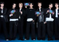 Members of BTS holding Oreo BTS packages, dressed in black and white outfits, smiling against a dark background.