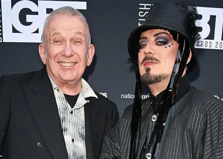 Jean Paul Gaultier and Boy George stand in front of a black wall with the logo 'British LGBT Awards' on it. Jean is wearing a striped white and grey shirt with a black blazer and is smiling. Boy George is wearing a pinstripe jacket and a black bucket-style hat with tassles. He has facial hair and gems around his eyes.