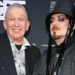 Jean Paul Gaultier and Boy George stand in front of a black wall with the logo 'British LGBT Awards' on it. Jean is wearing a striped white and grey shirt with a black blazer and is smiling. Boy George is wearing a pinstripe jacket and a black bucket-style hat with tassles. He has facial hair and gems around his eyes.