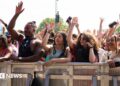 Crowd of festivalgoers standing behind a metal safety barrier at an outdoor music event, many with their hands raised in the air. A group at the front includes several young adults leaning on the barrier, some holding drinks and mobile phones, with trees and stage equipment visible in the background under bright daylight.