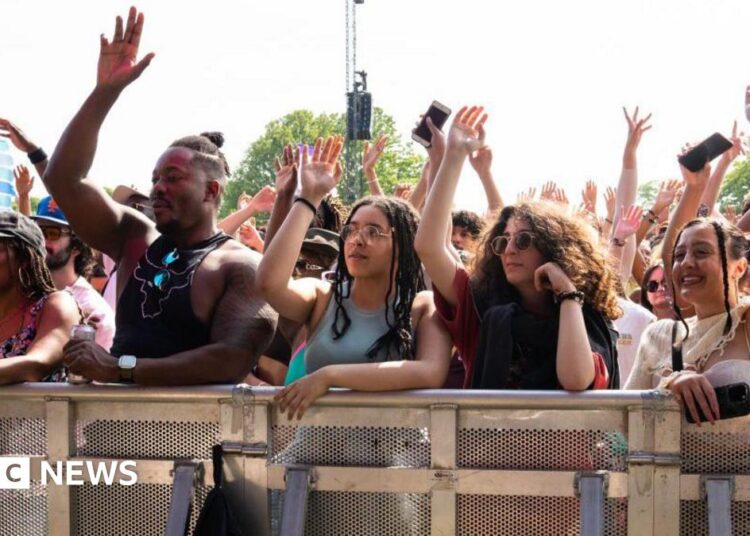 Crowd of festivalgoers standing behind a metal safety barrier at an outdoor music event, many with their hands raised in the air. A group at the front includes several young adults leaning on the barrier, some holding drinks and mobile phones, with trees and stage equipment visible in the background under bright daylight.